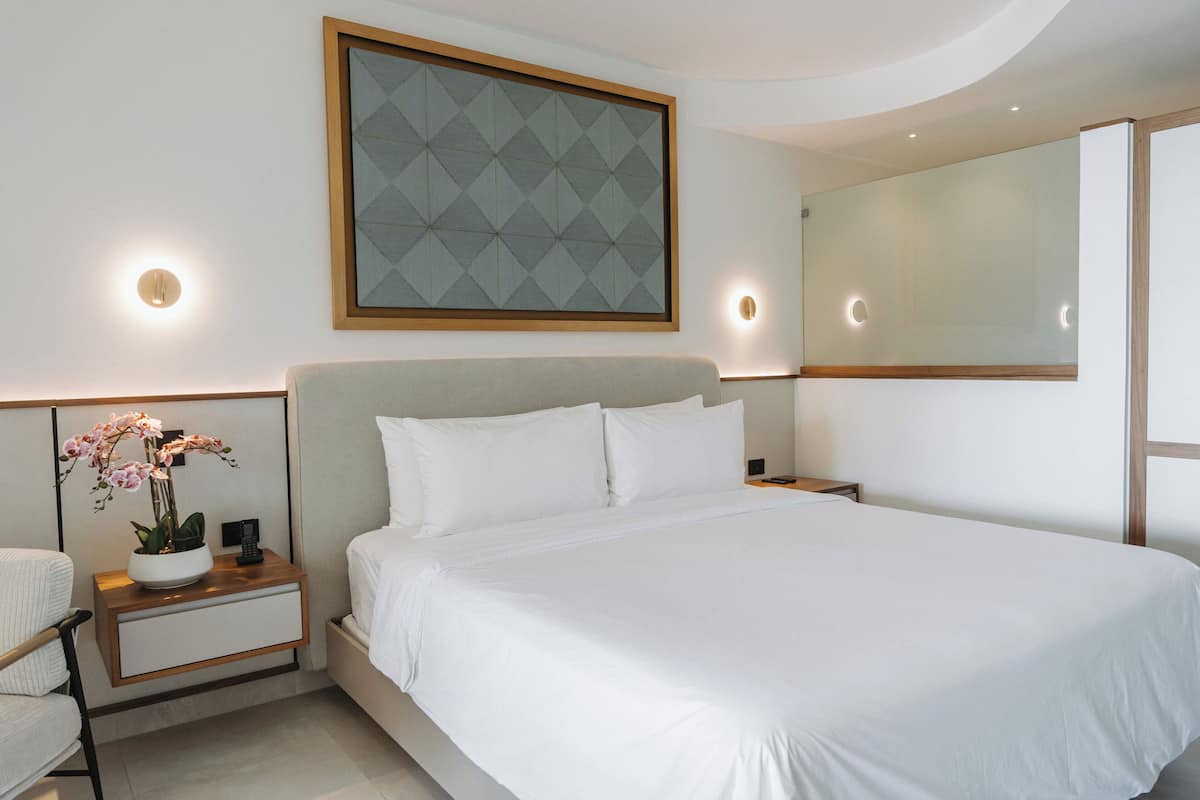 Una habitación de hotel moderna con una cama grande, una mesita de noche con un jarrón de flores, un espejo montado en la pared y una obra de arte con un diseño geométrico.