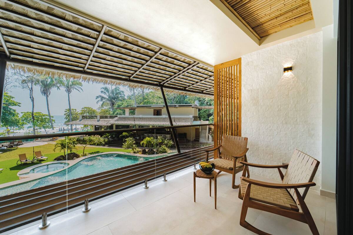 Un balcón con vistas a una piscina y un paisaje tropical, muebles de madera y un muro de piedra.