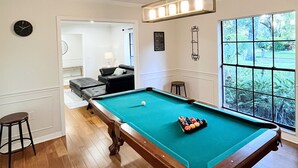 A pool table in a room with hardwood floors, a clock on the wall, and a window with a view of greenery. - Shady Oaks Estate near Tampa (Brandon)