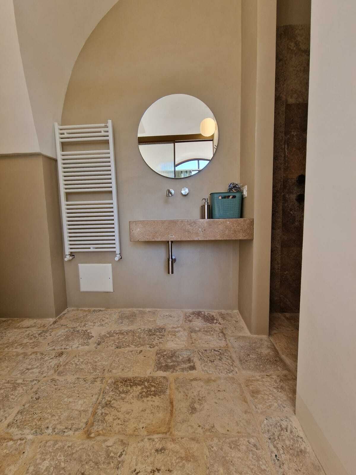 A bathroom with a stone-tiled floor, a round mirror, a white towel rack, a sink with a green soap dish, and a towel dispenser.