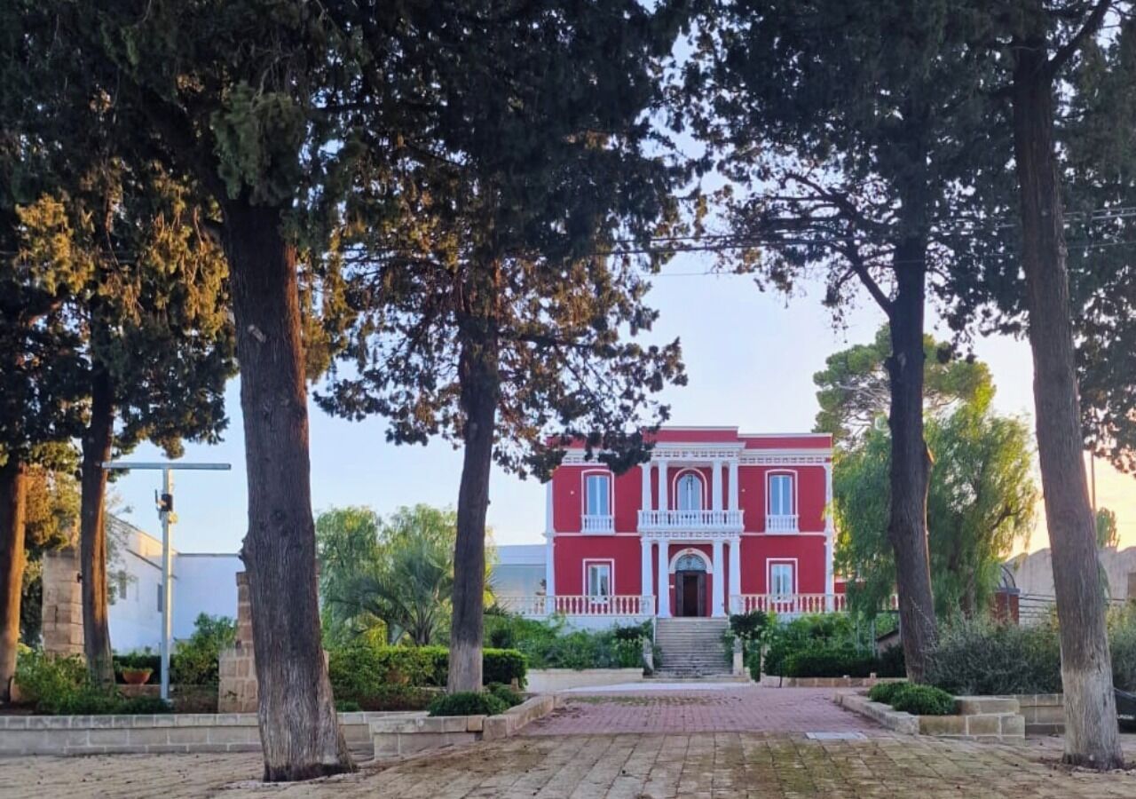 Un bâtiment rouge avec un balcon blanc, entouré d’arbres et bordé d’une allée pavée.