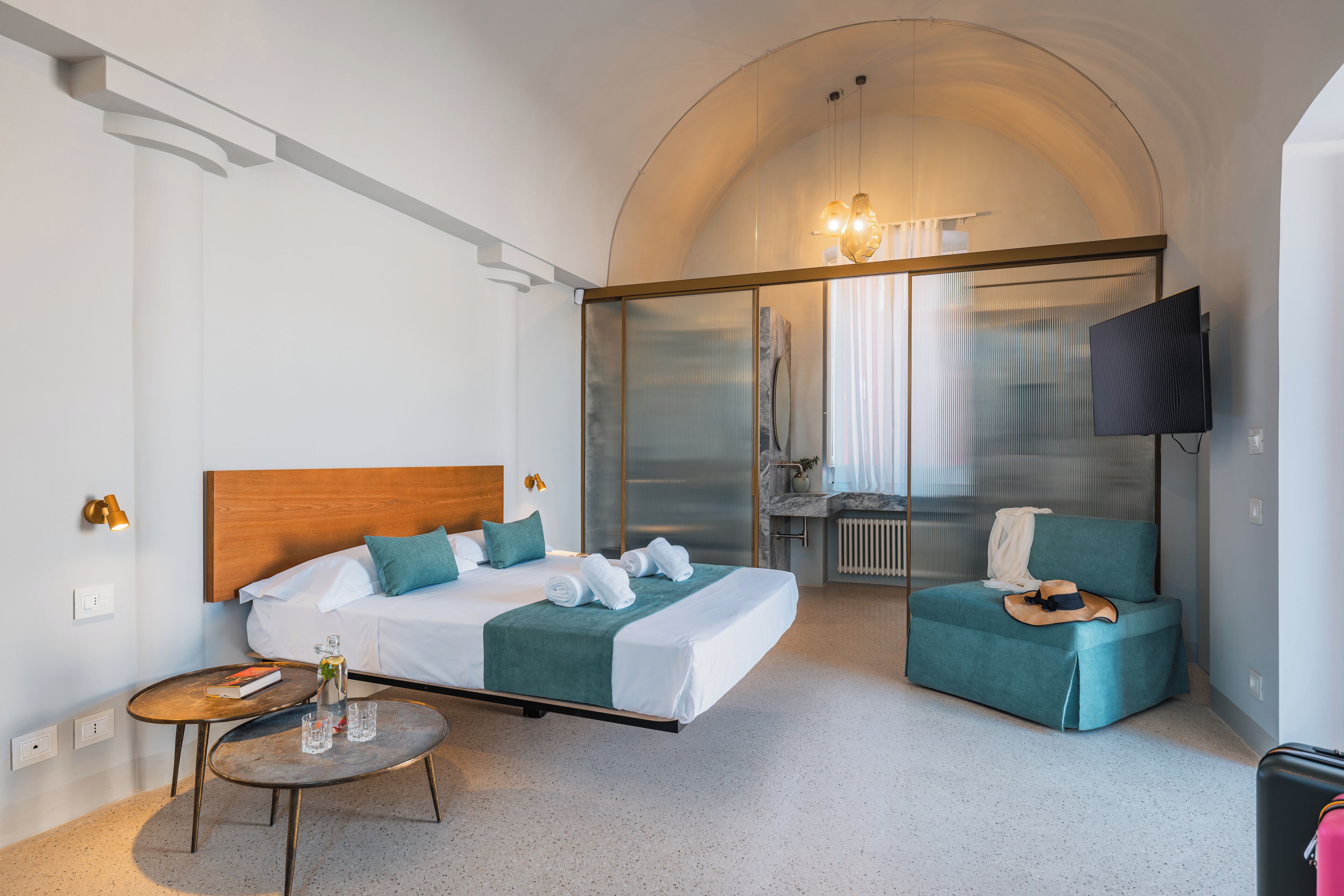 Una habitación de hotel moderna con una cama, zona de estar, televisor y baño visible a través de una puerta de cristal.