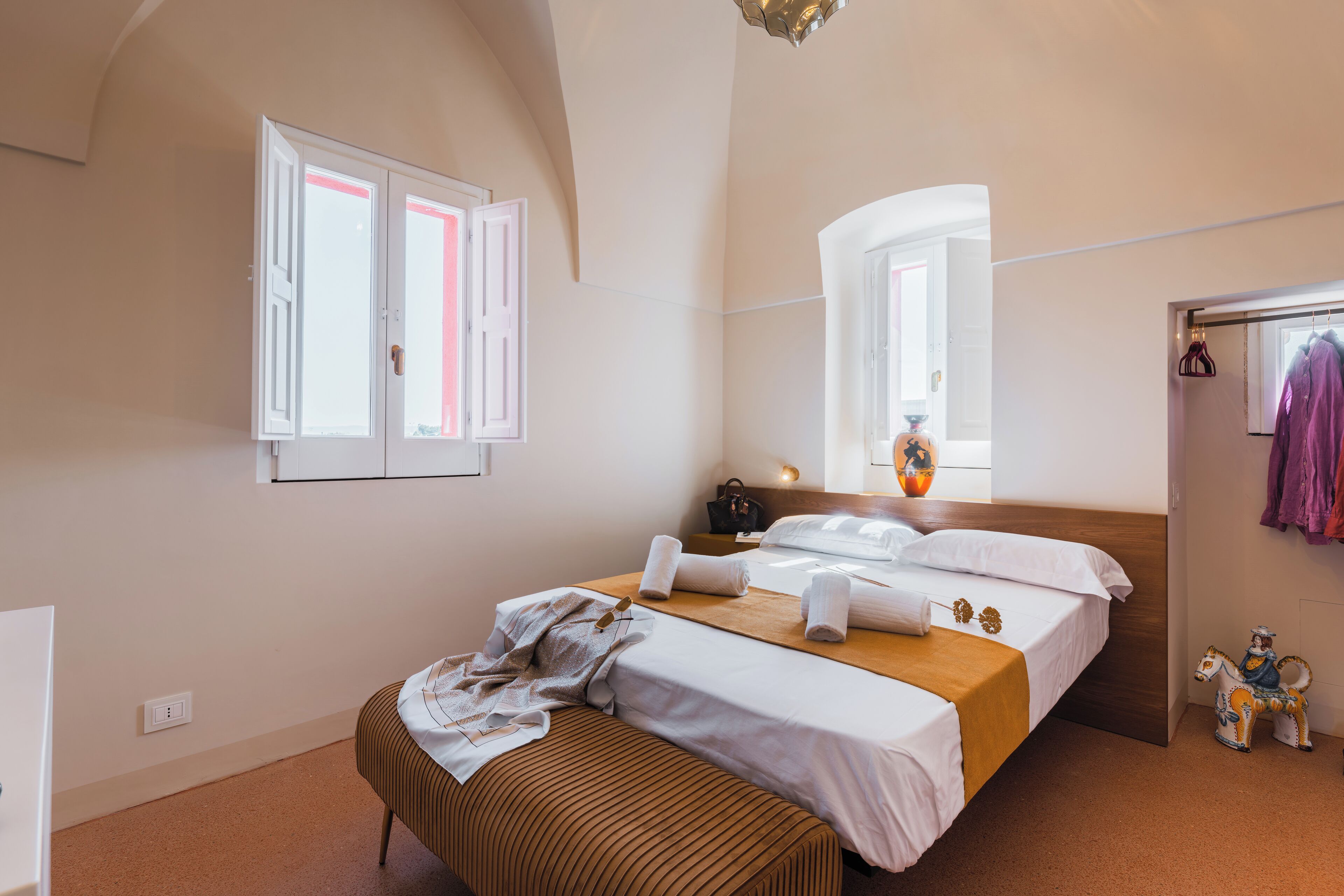 Quarto com cama, um banquinho, espelho, janela e um cabideiro com roupas.