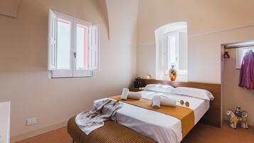 Quarto com cama, um banquinho, espelho, janela e um cabideiro com roupas.