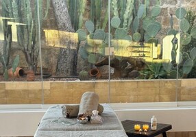 Un'area benessere con lettino da massaggio, asciugamani, candele e vista sui cactus attraverso una grande finestra.