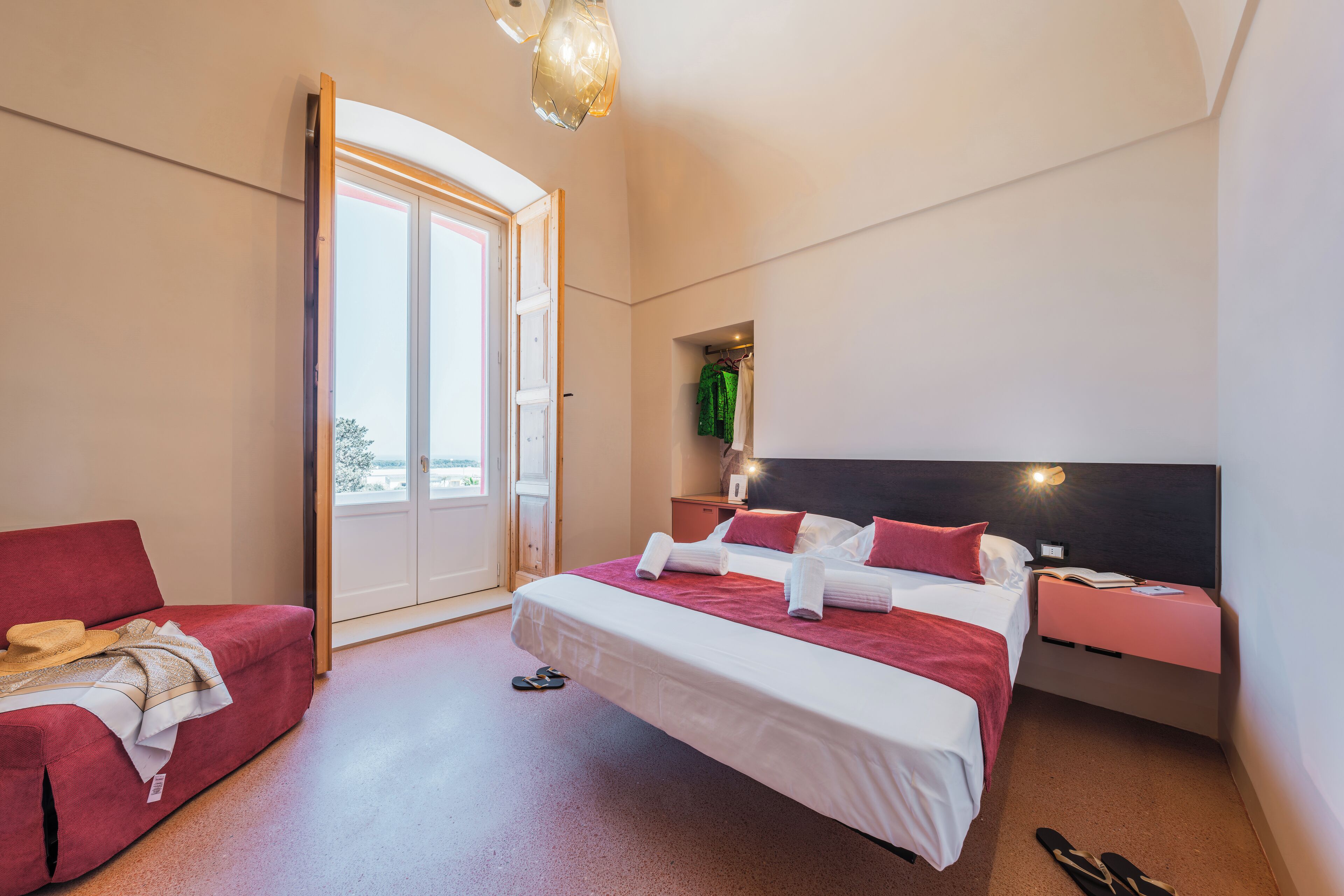 A modern hotel room with a bed, a red sofa, a nightstand, and a large window.