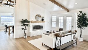 A spacious living room with a fireplace, white walls, wooden beams, and large windows. - Luxury Retreat with Spectacular Views (Draper)