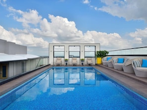 Una piscina en la azotea con vistas despejadas al horizonte de la ciudad, sillas de descanso y fachada de un edificio moderno.