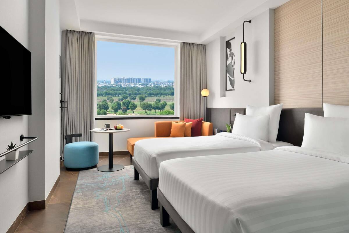 A hotel room with two beds, a TV, a small table with a cup and fruit, and a view of a cityscape.