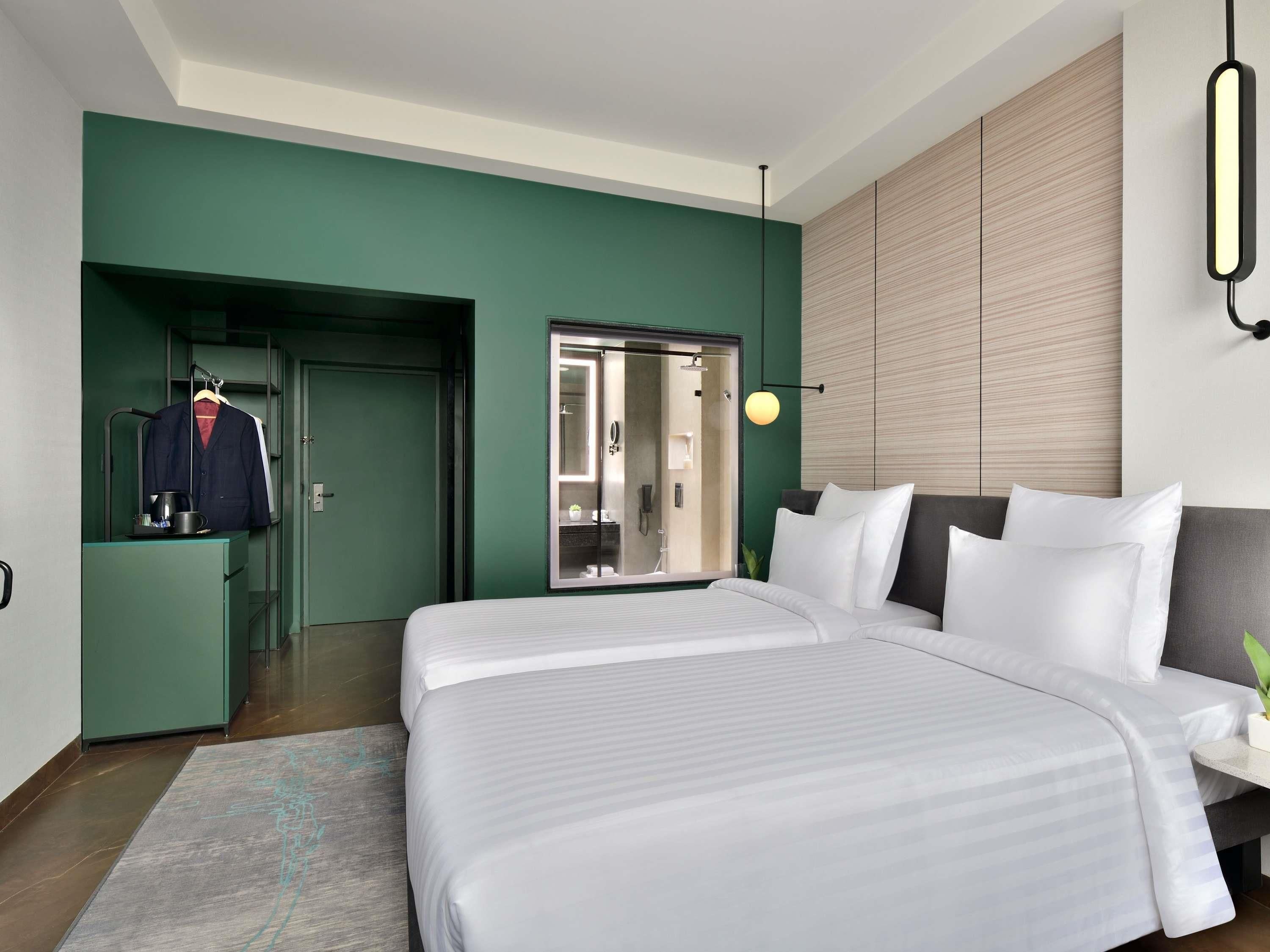 A modern hotel room with a large bed, a wardrobe, a mirror, and a bathroom visible through a glass door.