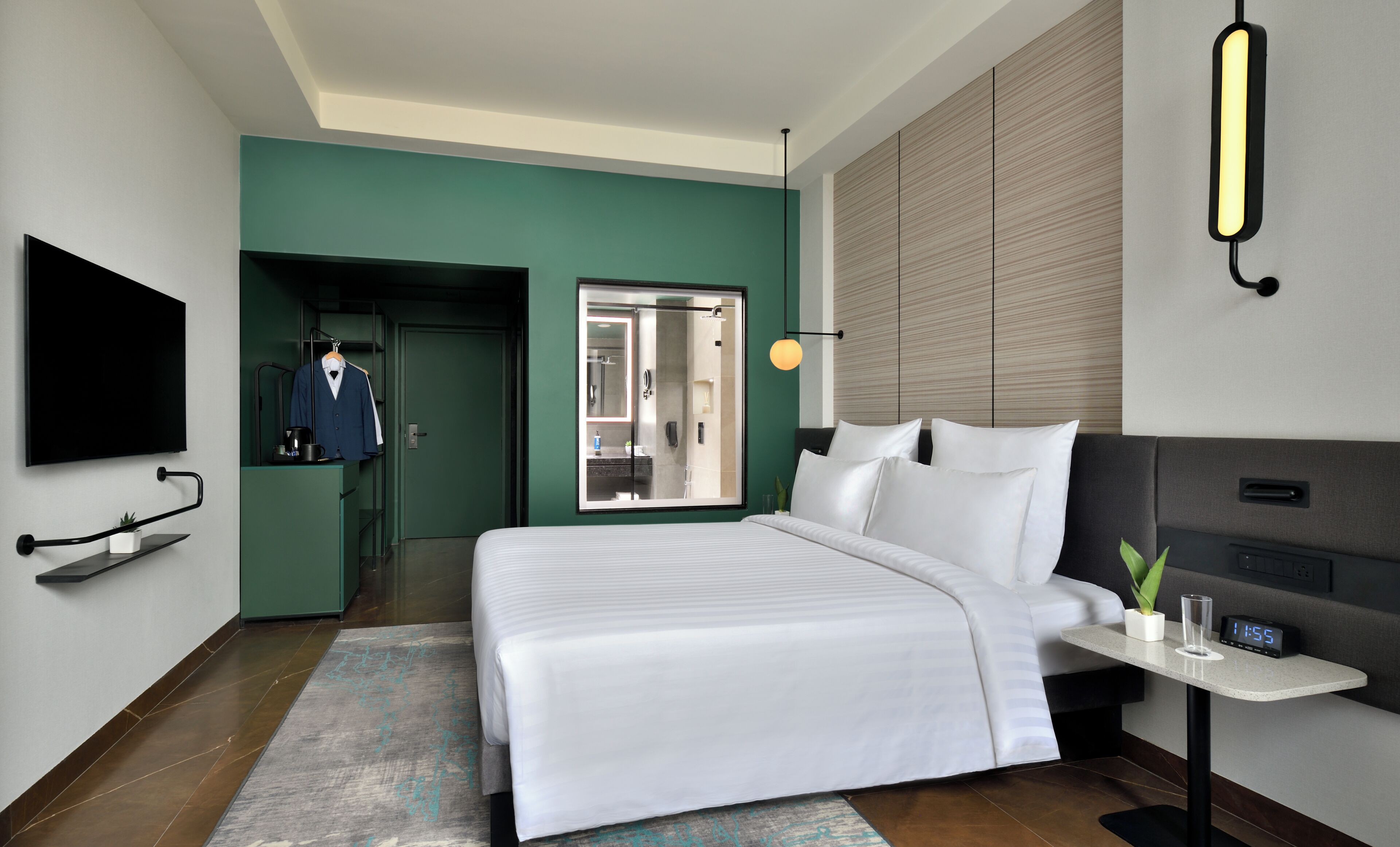 A modern hotel room with a large bed, a TV mounted on the wall, a desk with a clock, and a bathroom visible through an open door.