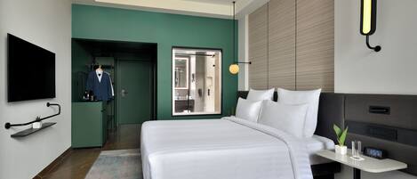 A modern hotel room with a large bed, a TV mounted on the wall, a desk with a clock, and a bathroom visible through an open door.