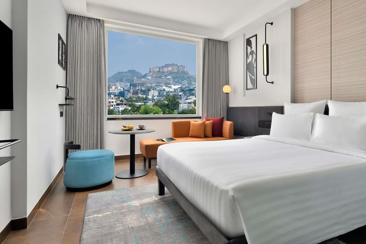 Une chambre dâhĂŽtel avec un grand lit, une petite table avec des fruits, une chaise bleue et une vue sur le paysage urbain.