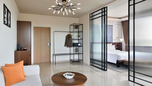 Ein modernes Hotelzimmer mit Sofa, einem kleinen Holztisch, einem Kleiderhaken an der Wand, einem Bett und einem großen Fenster mit Vorhängen.