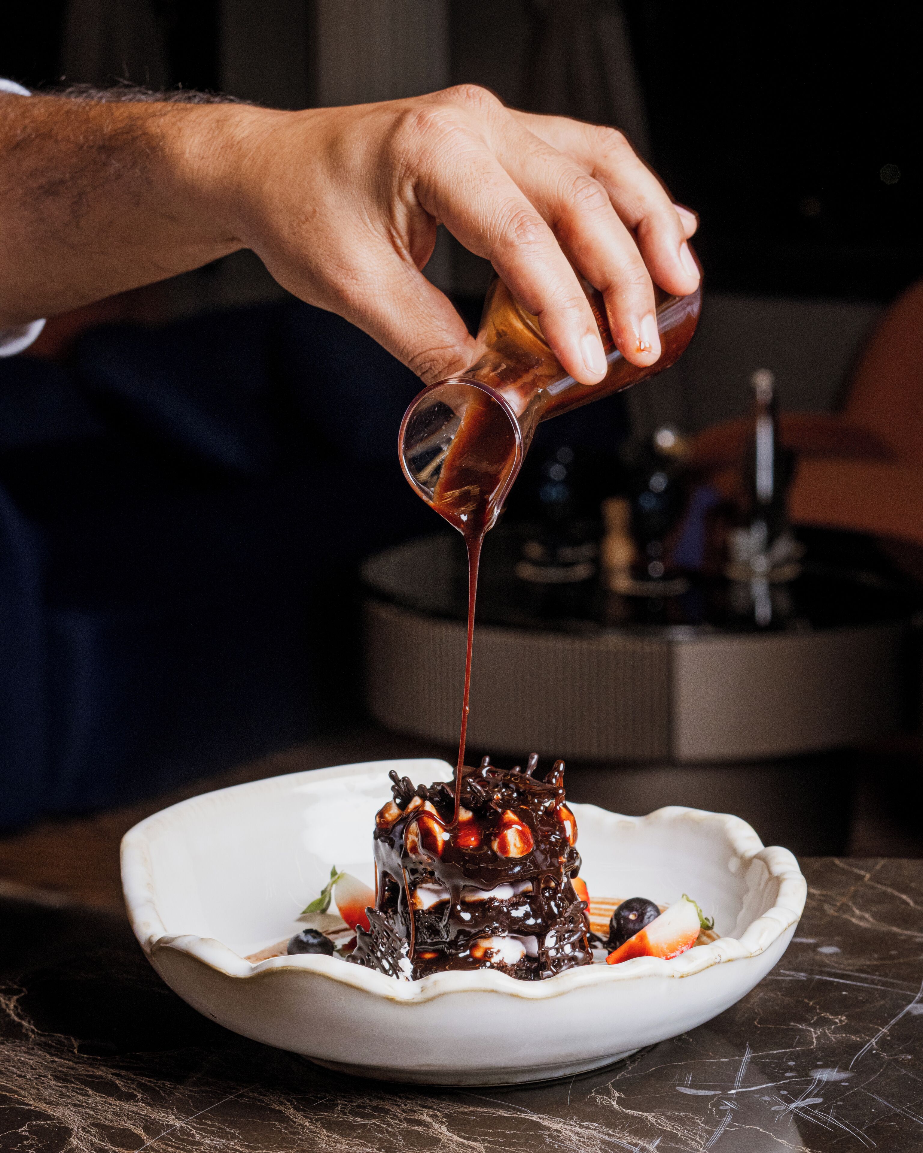 A person pouring a dark liquid onto a dessert with chocolate and fruit on a white plate.