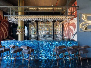 Una zona de bar con mostrador de mosaico azul, varias estanterías con botellas y taburetes de bar.