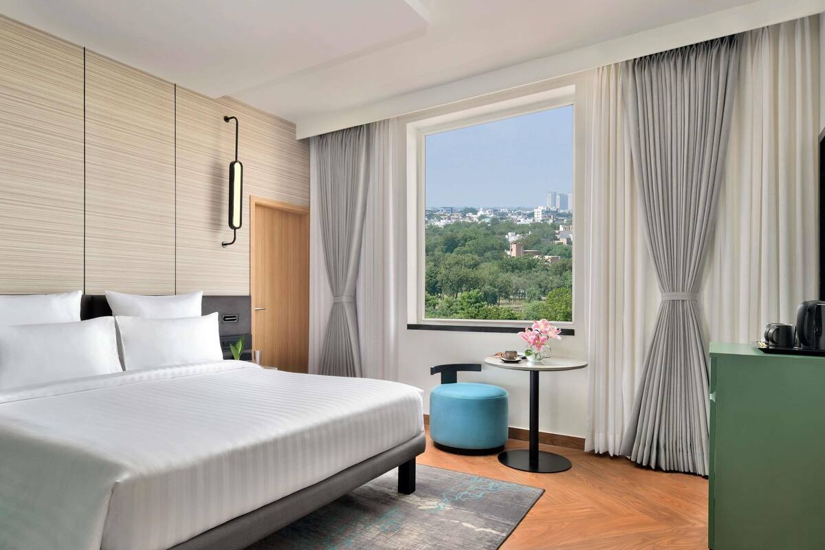 A hotel room with a large bed, a small table with a vase of flowers, a green cabinet, and a window with a city view.
