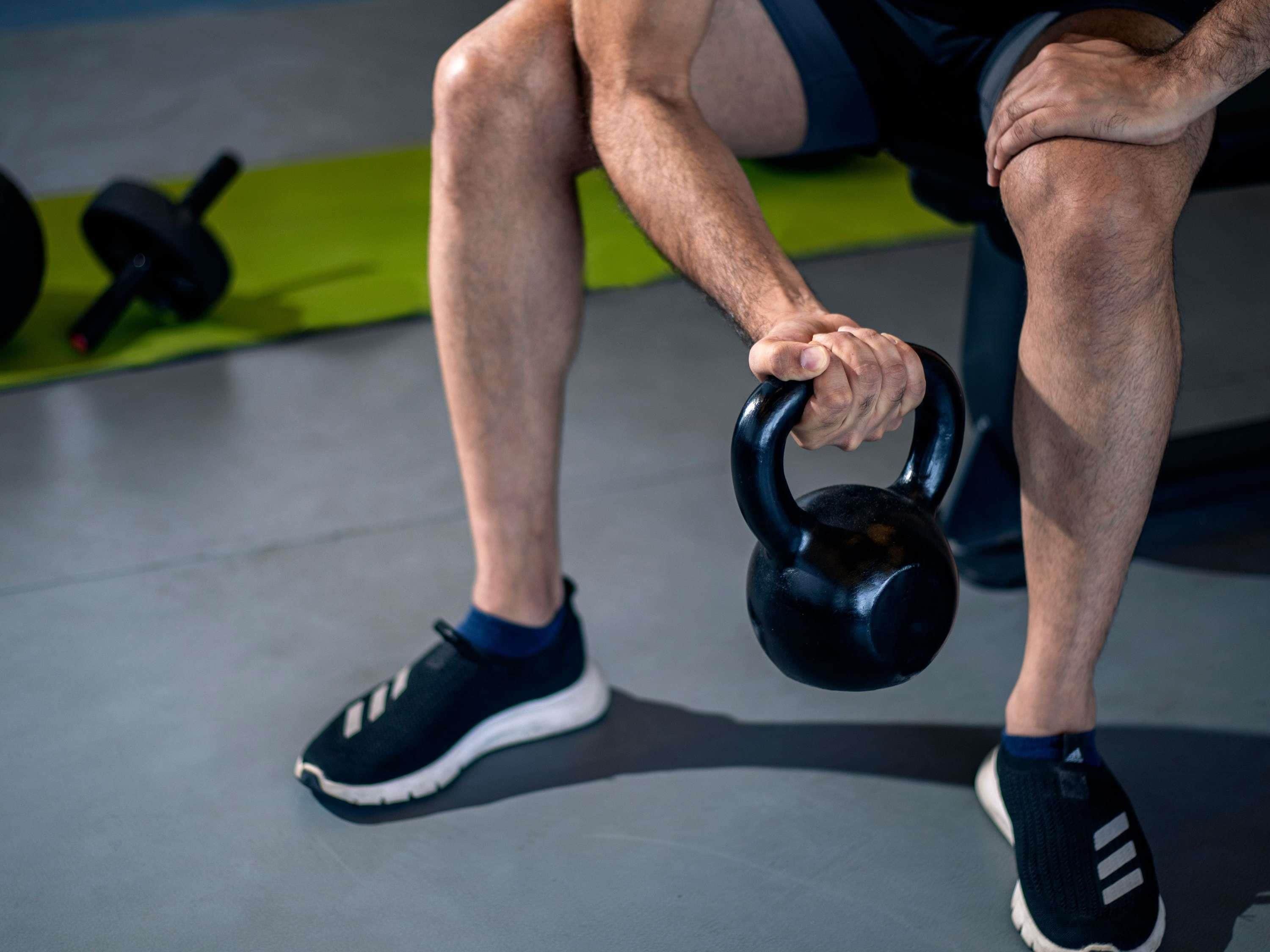 A person holding a kettlebell, wearing black and white sneakers, with gym equipment in the background.