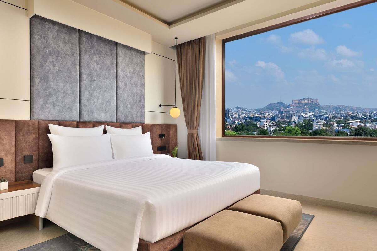 A modern hotel room with a large bed, a nightstand, a lamp, and a view of a cityscape.
