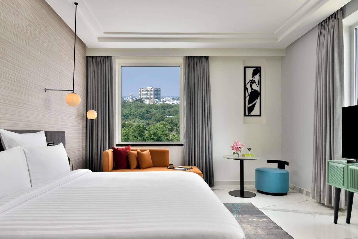 A modern hotel room with a large bed, a sofa, a TV, and a view of a cityscape.