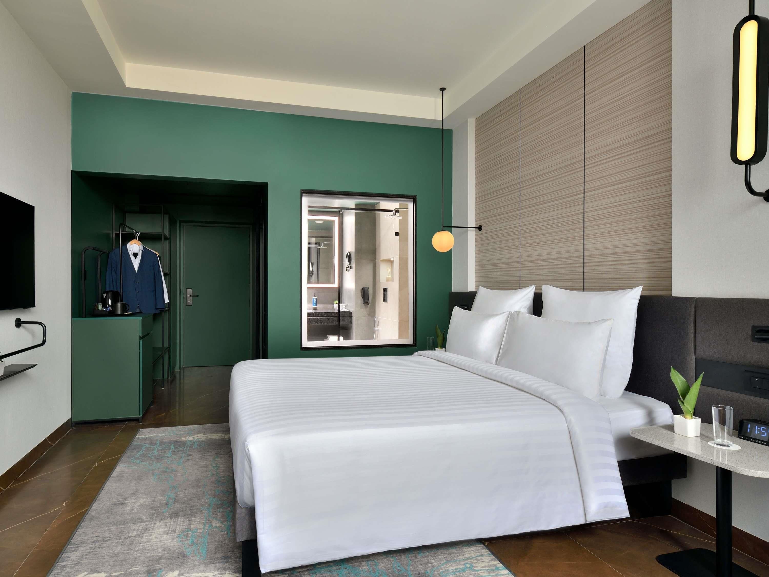 A modern hotel room with a large bed, a small bedside table with a clock, a TV mounted on the wall, and a mirror above the bed.