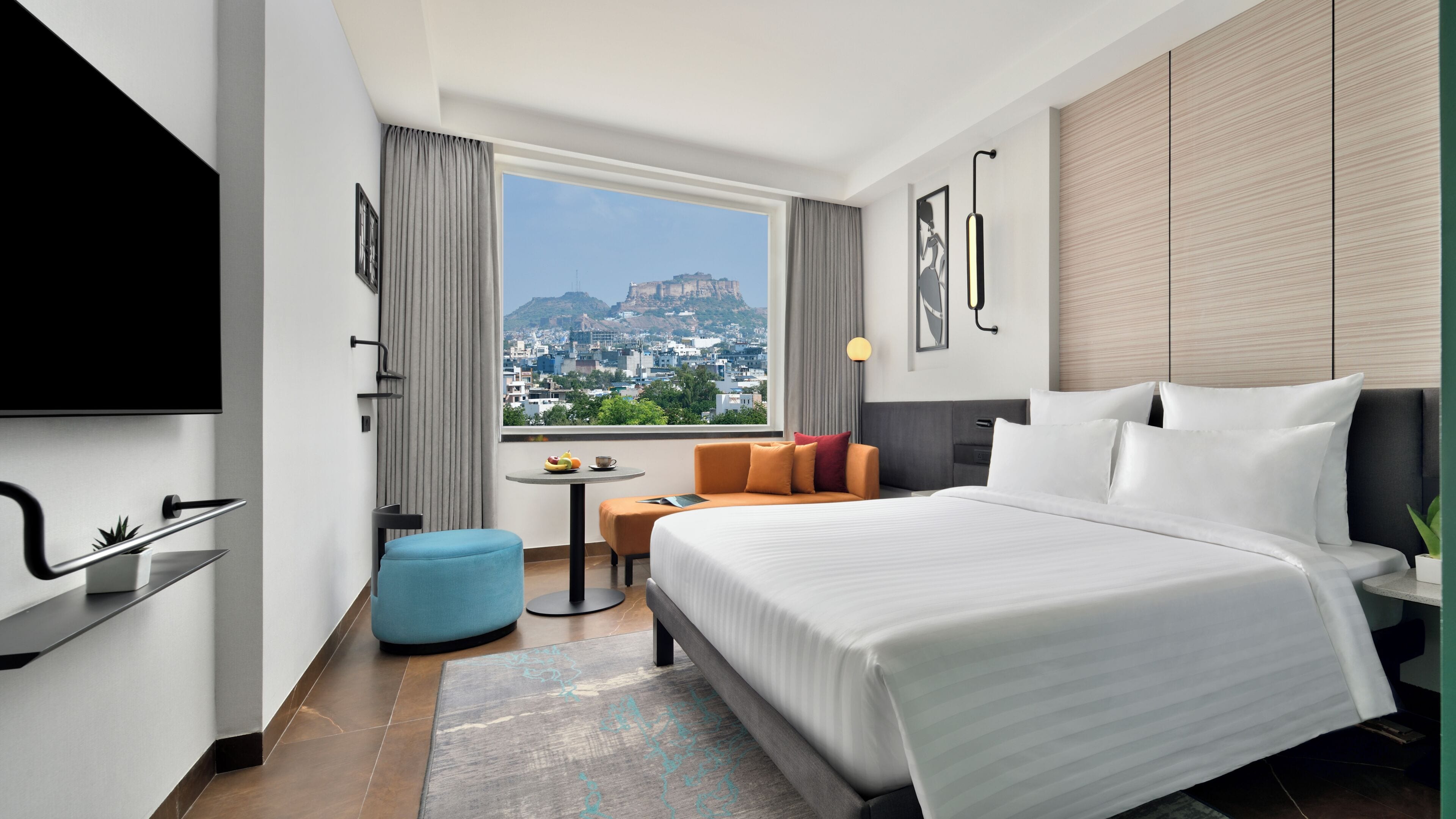 A hotel room with a bed, a sofa, a small table, and a view of a cityscape.