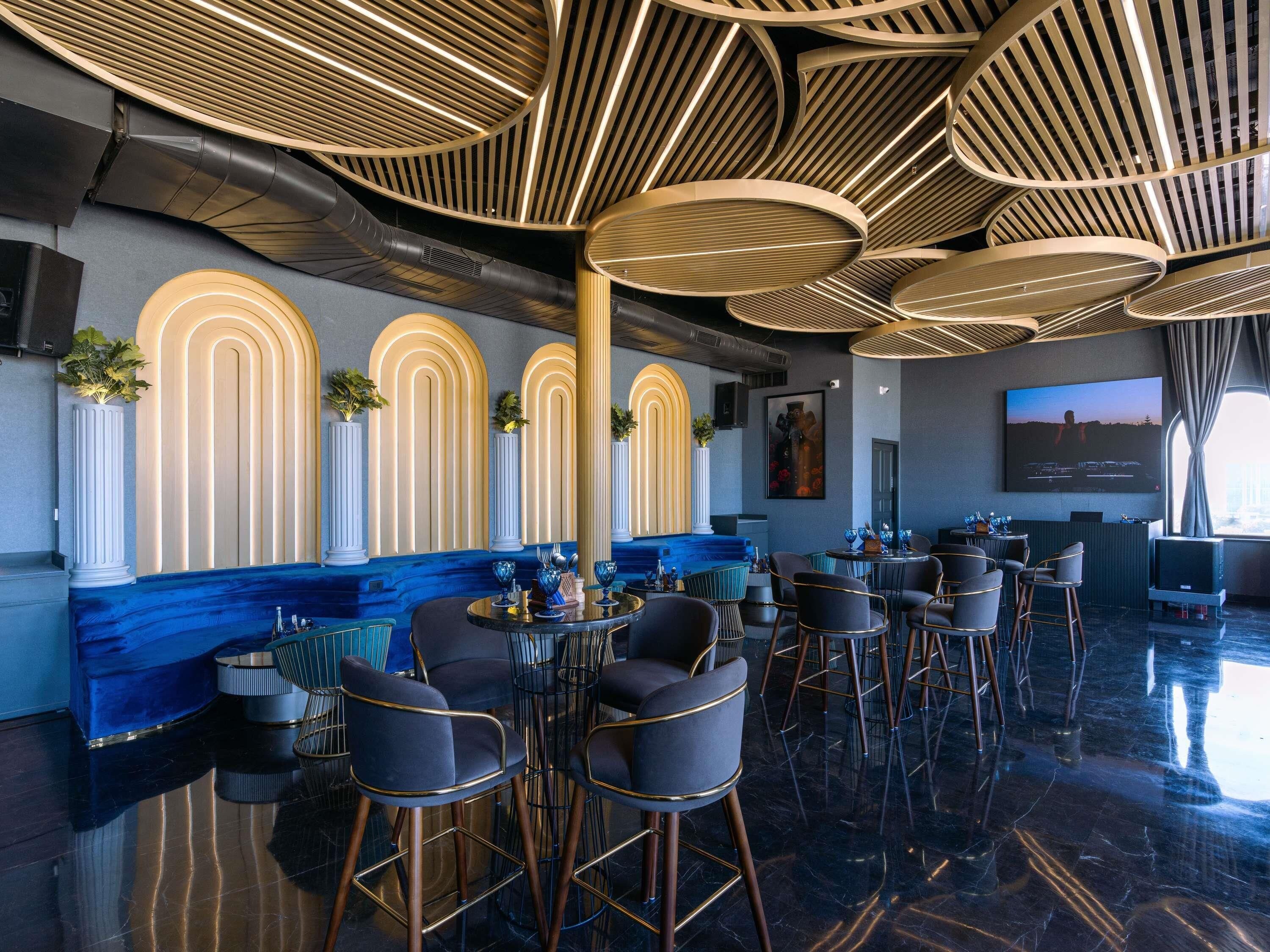 A modern bar area with high ceilings, circular light fixtures, and a seating arrangement with bar stools and tables.