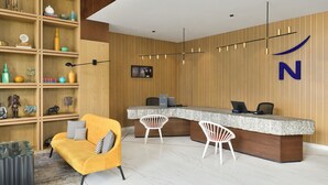 Uma receção com painel de madeira, duas cadeiras brancas, um sofá amarelo e uma estante com diversos objetos decorativos.