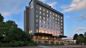 Um hotel de vários andares com um destaque para o letreiro 'NOVOTEL' na fachada, apresentando um design arquitetônico moderno que combina elementos de vidro e tijolo.
