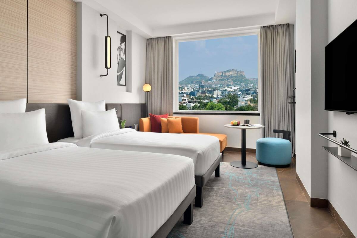 A hotel room with two beds, a small table, a blue ottoman, a TV, and a city view.