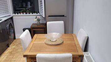 A compact kitchen with a wooden table, white chairs, a refrigerator, a window with blinds, and a radiator.