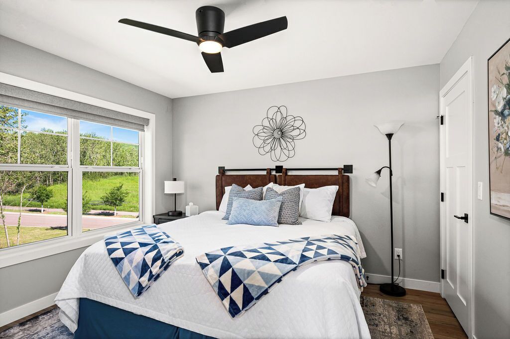 A bedroom with a bed, a ceiling fan, a window with a view, a floor lamp, and a wall art piece.