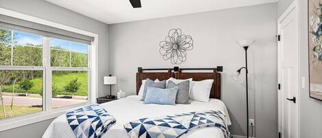 A bedroom with a bed, a ceiling fan, a window with a view, a floor lamp, and a wall art piece.