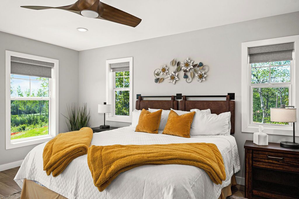 A bedroom with a large bed, two windows with views of greenery, a ceiling fan, and a wall-mounted decorative piece.
