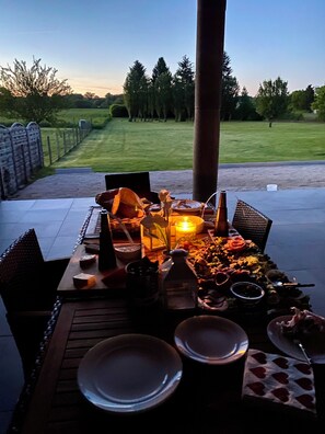 An outdoor dining area with a table set for a meal, surrounded by chairs, and a view of a grassy field and trees in the background.