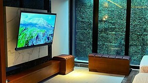 A modern living room with a large flat-screen TV mounted on a wooden console, a cushioned bench, and a large window offering a view of greenery. - Calle 76 Estudio by Caribecheap (Barranquilla)