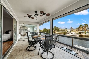 A balcony with a table, chairs, and a ceiling fan. It overlooks a body of water with boats and houses in the background. - Waterfront home w/ direct gulf access + pool + hot tub + activites! (Hernando Beach)