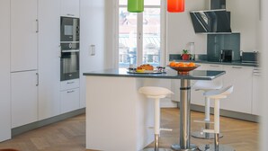 A modern kitchen with white cabinetry, a central island with stools, a built-in oven, and a window with a view of buildings. - Bright, quiet apartment in the heart of Biarritz (Biarritz)