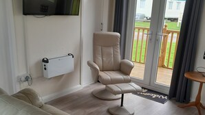 Ett modernt vardagsrum med en beige soffa, en fåtölj som kan lutas bakåt, en platt-TV som är monterad på väggen och utsikt över utomhusmiljön genom en skjutdörr i glas.