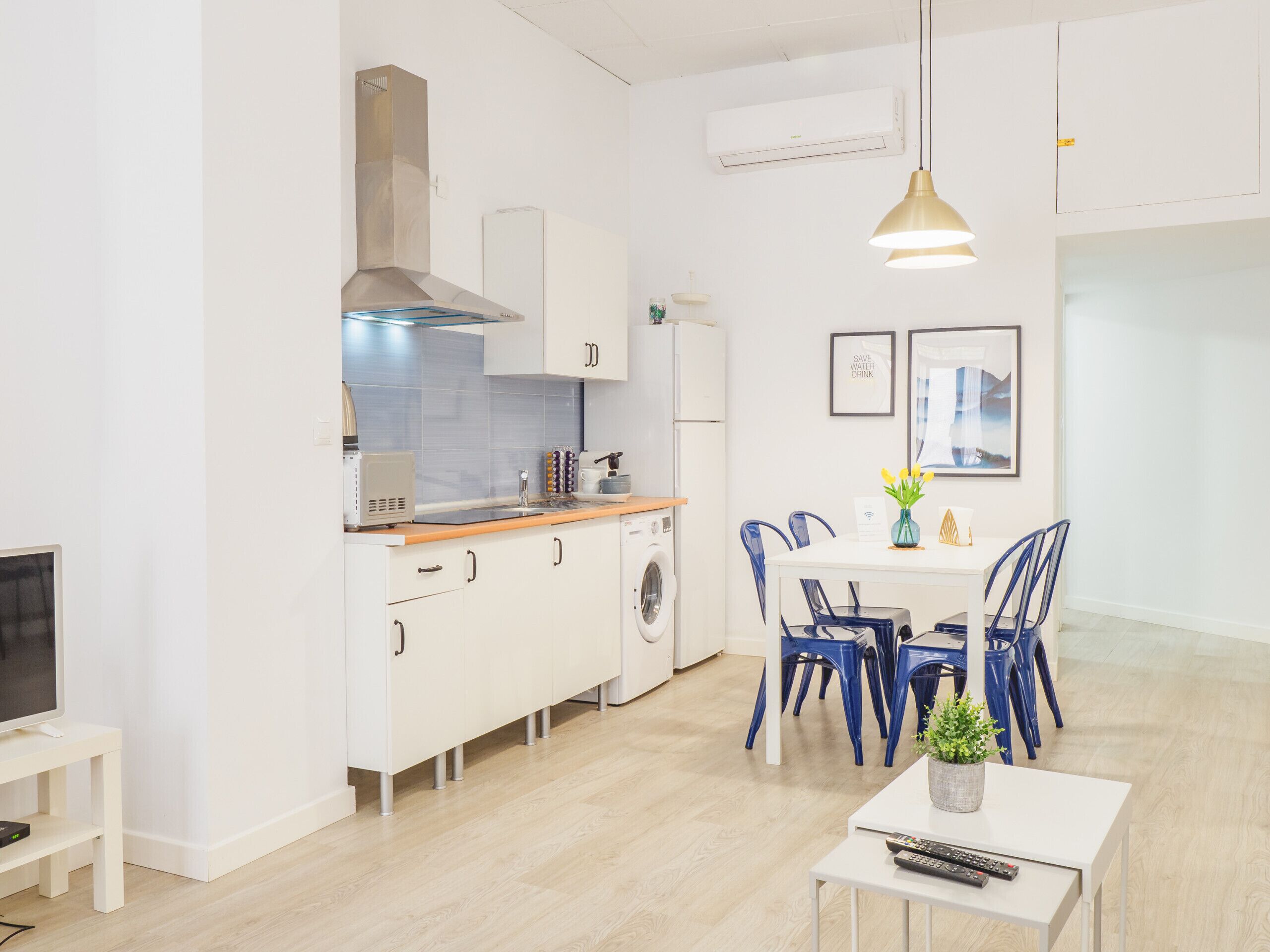 Una cocina moderna con armarios blancos, horno empotrado y microondas. Hay una zona de comedor con una mesa blanca y sillas azules.
