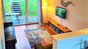 A modern living room with a large sliding glass door leading to a patio, a flat-screen TV mounted on the wall, and a small kitchen area with a sink. - Cornish Getaway (Newquay)