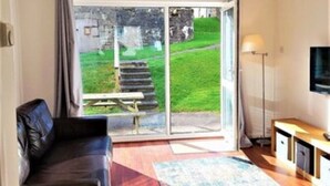 A modern living room with a sofa, a coffee table, and a TV on a stand. There is a sliding glass door leading to a garden with a bench and a lamp. - Cornish Getaway (Newquay)