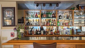 Una zona de bar con estantes llenos de botellas de licor y vasos, una barra de madera y un detector de incendios en la pared.