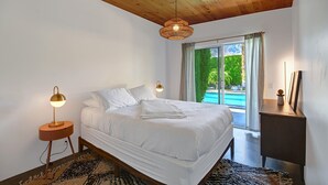 Une chambre avec un grand lit, un plafond en bois, une table de chevet avec une lampe, un bureau et une vue sur une piscine.