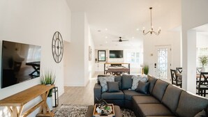 A modern living room with a sectional sofa, a flat-screen TV, a wooden bench, and a decorative clock. - Palmeras- Pool, Views, Game Room (Indian Wells)