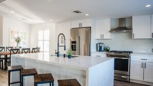 A modern kitchen with white cabinets, a large island with stools, a stainless steel refrigerator, and a gas stove. - Palmeras- Pool, Views, Game Room (Indian Wells)