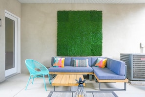 Uma área de estar moderna ao ar livre, com um sofá azul, almofadas coloridas, uma cadeira turquesa e uma mesa de madeira.