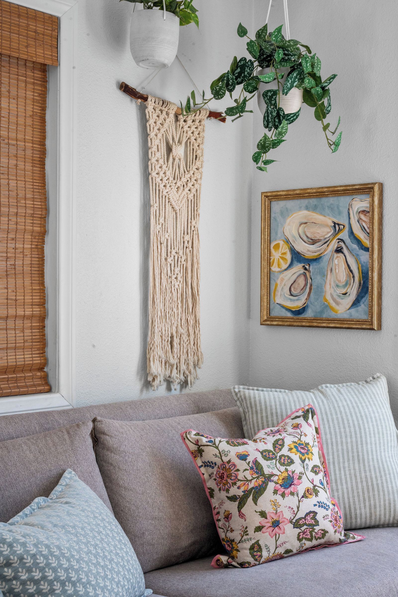 A living room with a macrame wall hanging, a hanging plant, a framed painting, and a sofa with patterned cushions.
