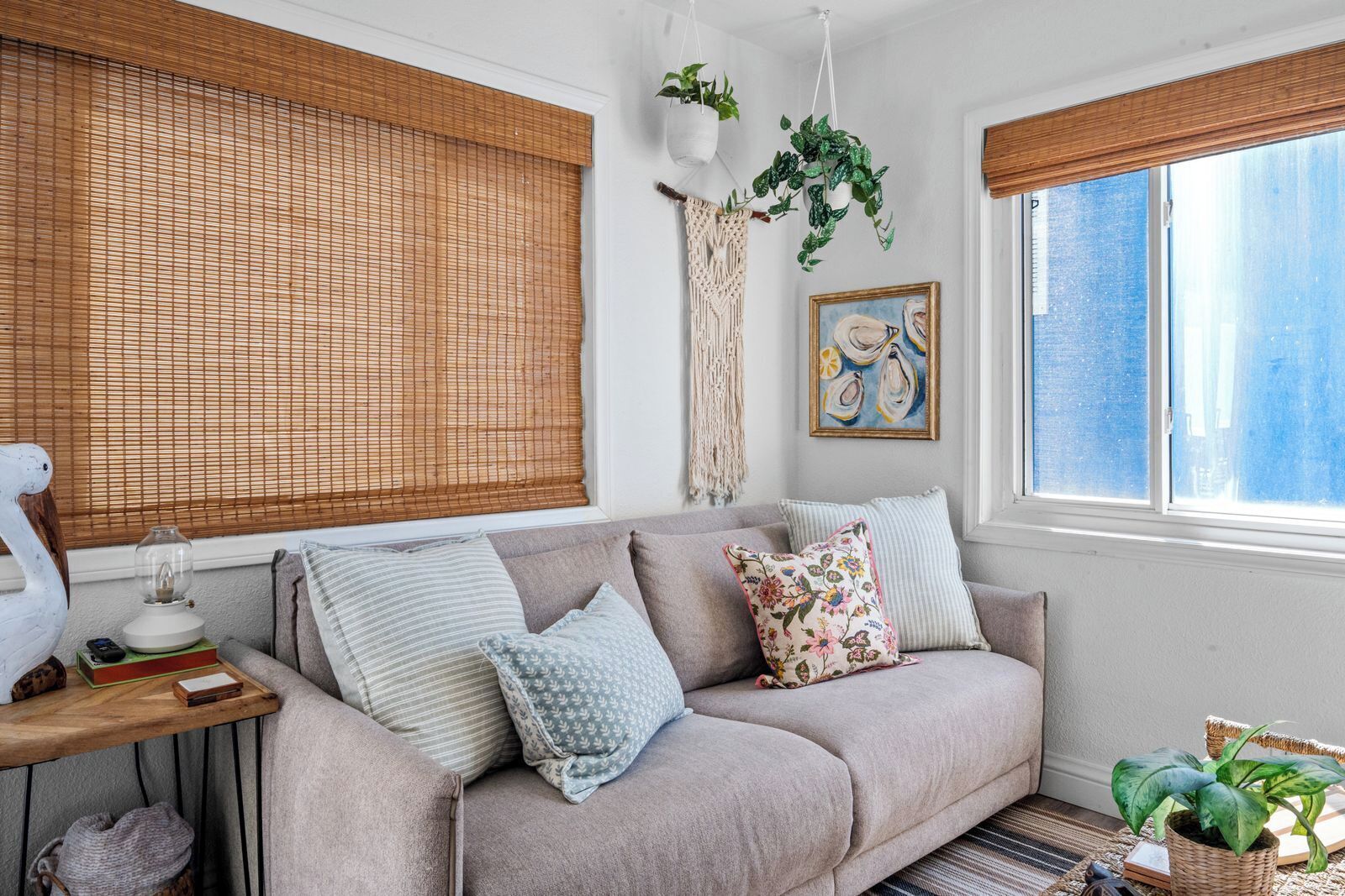 A cozy living room with a sofa, cushions, a lamp, a macrame wall hanging, a framed artwork, and a window with a view.