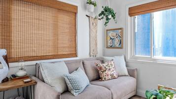A cozy living room with a sofa, cushions, a lamp, a macrame wall hanging, a framed artwork, and a window with a view.
